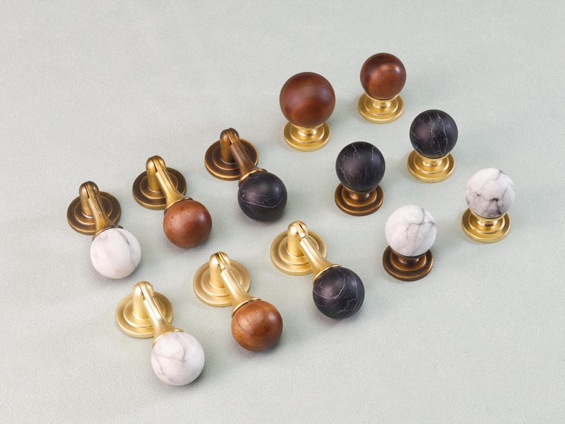 May include: A collection of cabinet knobs with brass bases and various tops. The tops include wood, marble, and black marble. The knobs are arranged on a light green surface, showcasing their different styles and finishes.
