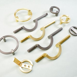 May include: An assortment of cabinet hardware in various designs and finishes. The collection includes ring pulls, curved handles, and circular knobs in gold and gray tones. The hardware is arranged on a light-colored surface, showcasing the different styles.