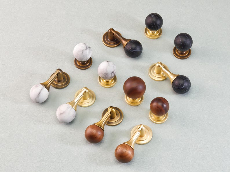 May include: An assortment of cabinet knobs and pulls in various styles. Some have marble-like spheres, others are wood, and some are black. All have brass or bronze-colored bases and stems. The knobs and pulls are arranged on a light gray surface.