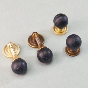 May include: A collection of cabinet hardware featuring black marble-effect knobs and pulls with brass and gold-toned bases. The hardware includes both round knobs and pulls with a decorative design.