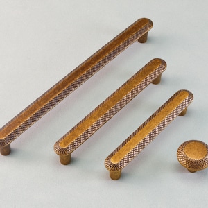 Puede incluir: Un juego de cuatro piezas de herrajes para gabinetes de color bronce. El juego incluye tres tiradores de barra de diferentes tamaños y un pomo redondo. Cada pieza tiene un patrón texturizado, entrecruzado y extremos redondeados.
