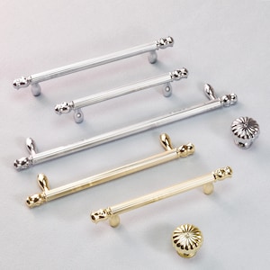 May include: An assortment of silver and gold cabinet hardware, including several bar pulls and two round knobs. The pulls have a ribbed design and decorative ends. The knobs have a fluted design.