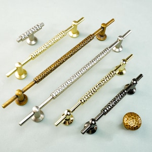 May include: A collection of decorative cabinet hardware in various metallic finishes. The assortment includes several pull handles with textured shafts and knob hardware. Finishes include gold, silver, bronze, and black.