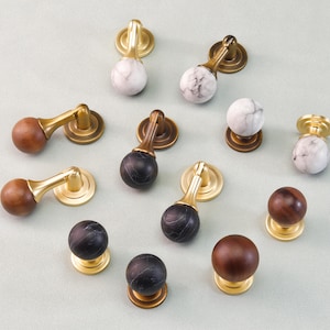 Może przedstawiać: Asortyment gałek i uchwytów do szafek w różnych wzorach. Okucia posiadają elementy mosiężne, drewniane i marmurowe. Gałki i uchwyty są w odcieniach brązu, czerni, bieli i złota. Okucia przeznaczone są do mebli i szafek.