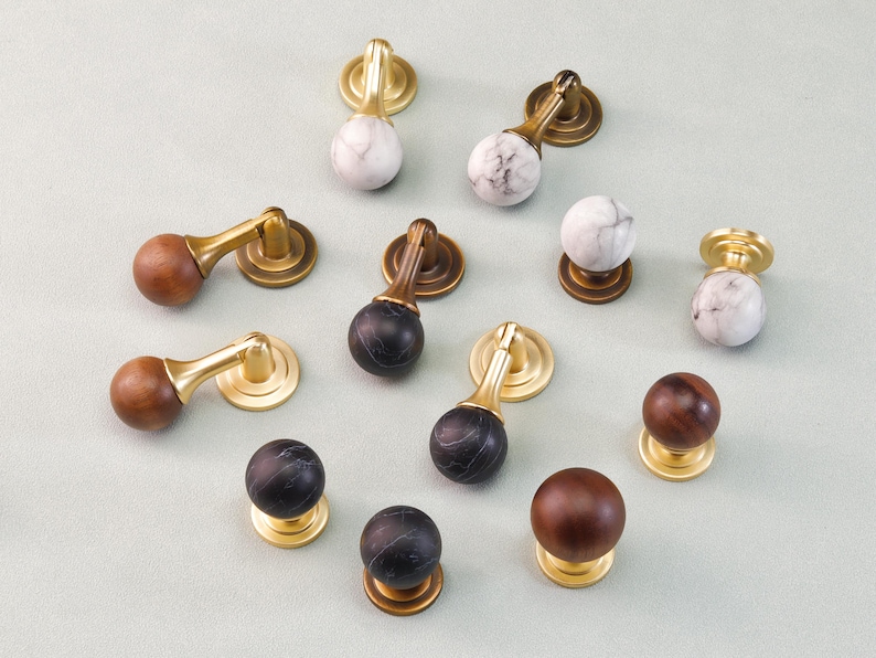 May include: An assortment of cabinet knobs and pulls. The knobs feature a variety of materials, including wood, marble, and dark stone, with brass or gold-toned hardware. The pulls have a similar design, with a curved handle and a round base.