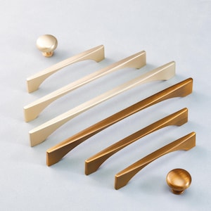 May include: A collection of gold and bronze cabinet hardware, including handles and knobs. The handles have a sleek, modern design with a curved shape. The knobs are round and have a matching finish. Ideal for kitchen or bathroom cabinets.