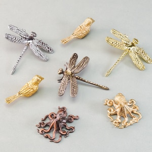 May include: A selection of decorative metal pins featuring insect and marine life designs. The pins include dragonflies, birds, and an octopus, in silver, gold, and bronze finishes. Intricate details are visible. The items are displayed on a light grey surface.