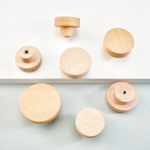May include: A collection of wooden cabinet knobs in various shapes and sizes. The knobs are made of light-colored wood and are arranged on a white and light blue surface. The knobs include round, cylindrical, and tiered designs.
