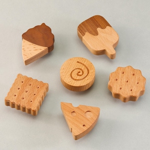 Könnte beinhalten: Eine Sammlung von sechs Holzspielzeugen in Form von Lebensmitteln. Das Set enthält eine Eistüte, ein Eis am Stiel, einen Keks, ein Käse-Eckstück, einen Cracker und ein rundes Stück mit Wirbelmuster. Die Spielzeuge sind aus hellem und dunklem braunem Holz.