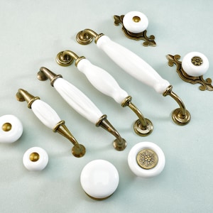 May include: A collection of white ceramic and brass cabinet hardware, including handles and knobs. The handles have a curved design, and the knobs feature a variety of brass accents. The hardware is arranged on a light blue surface.
