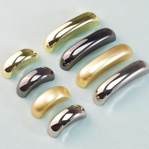 Könnte beinhalten: Eine Auswahl an gebogenen Schrankgriffen in verschiedenen Metallausführungen, darunter Gold, Silber und Schwarz. Die Hardware ist auf einer hellblauen Oberfläche angeordnet, die ihre polierten und reflektierenden Oberflächen hervorhebt. Diese Griffe sind für Möbel.