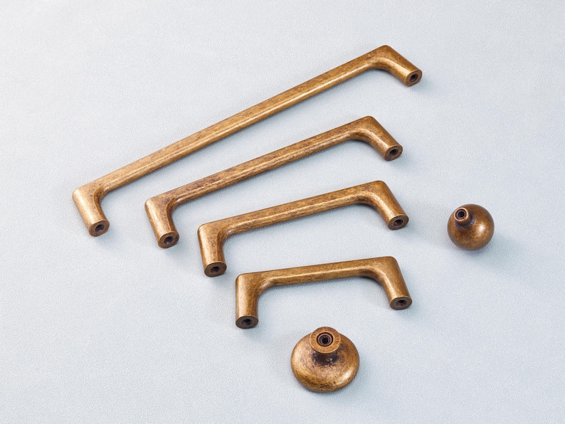 May include: A set of antique brass cabinet hardware, comprising four pull handles and two knobs. The handles are of varying lengths, with a curved design and mounting holes. The knobs are spherical and flat-based. The hardware has a warm, aged finish.