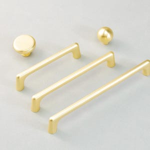 May include: A selection of gold-coloured cabinet hardware, featuring three bar pulls and two round knobs. The pulls have a simple, modern design, and the knobs are spherical. The hardware is arranged on a light grey surface.