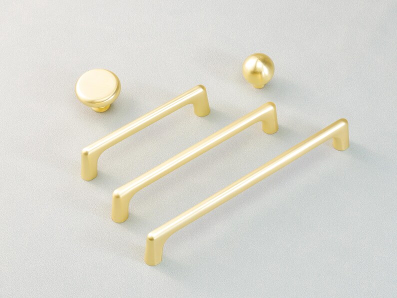 May include: A selection of gold-coloured cabinet hardware, featuring three bar pulls and two round knobs. The pulls have a simple, modern design, and the knobs are spherical. The hardware is arranged on a light grey surface.
