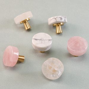 May include: A collection of decorative cabinet knobs in various colors and patterns. The knobs feature gold-colored stems and tops made of white, pink, and clear stones. The knobs are round and cylindrical, suitable for furniture or decor.