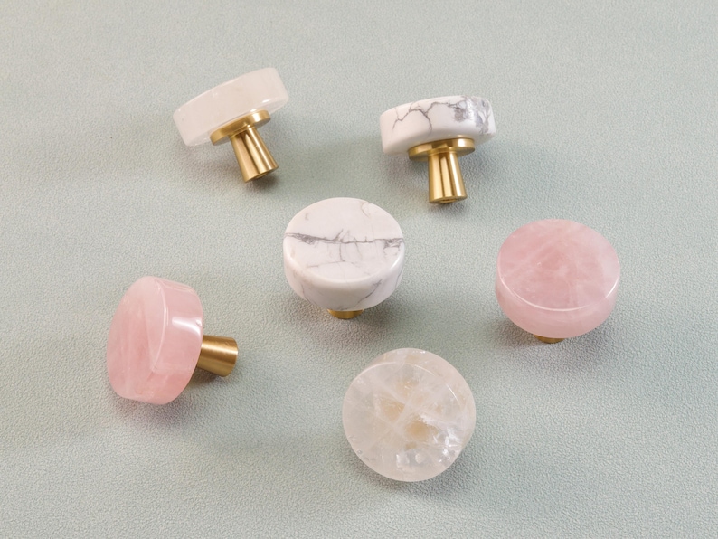 May include: A collection of decorative cabinet knobs in various colors and patterns. The knobs feature gold-colored stems and tops made of white, pink, and clear stones. The knobs are round and cylindrical, suitable for furniture or decor.