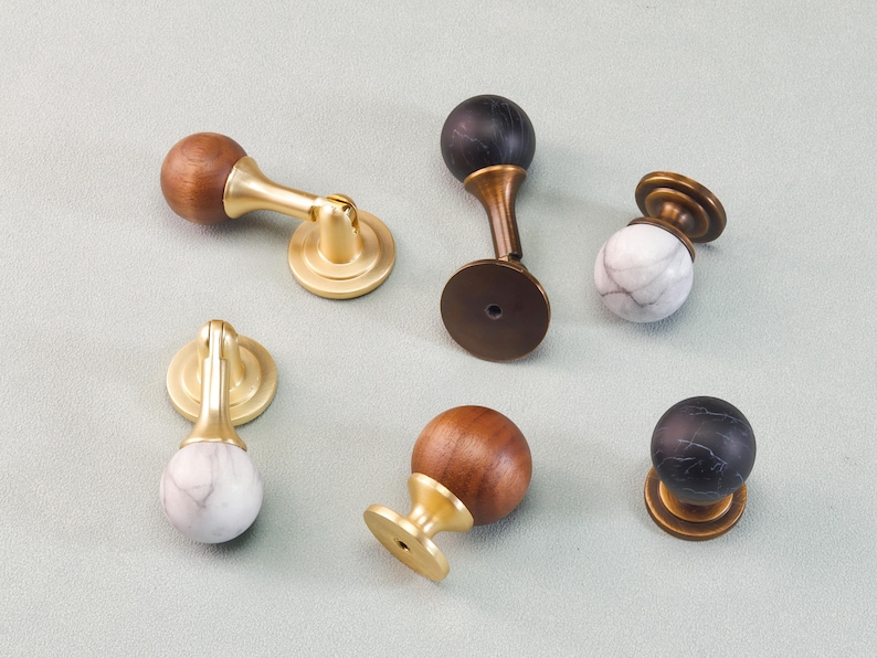 May include: A collection of cabinet knobs and pulls in various designs. Some feature a wooden sphere with a brass base, others have a marble sphere with a brass base, and some have a black marble sphere with a brass base.