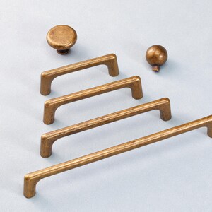 May include: A selection of antique brass cabinet hardware, comprising two knobs and three bar pulls. The knobs are round, and the pulls are rectangular with rounded ends. The hardware is presented against a light blue backdrop.