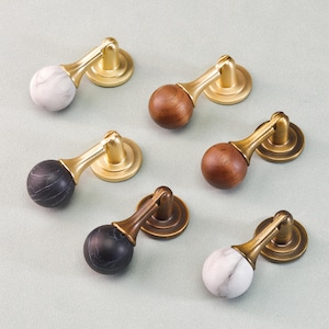 May include: A collection of cabinet pulls with brass hardware. The pulls feature marble, wood, and black marble spheres. The hardware is a warm gold color. The pulls are arranged on a light gray surface.