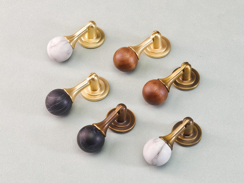 May include: A collection of cabinet pulls with brass hardware. The pulls feature marble, wood, and black marble spheres. The hardware is a warm gold color. The pulls are arranged on a light gray surface.