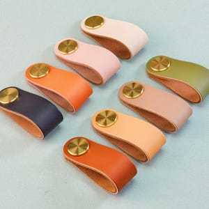 May include: An assortment of leather cabinet pulls in various colors, including pink, orange, navy, and green. Each pull features a gold-colored circular accent. The pulls are arranged on a light blue surface.
