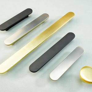 May include: An assortment of modern cabinet hardware, including long bar pulls in black, brushed nickel, and gold finishes. A gold knob is also included. The hardware is arranged on a light blue surface.