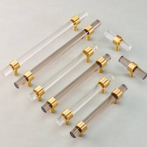 May include: A collection of clear and smoky gray acrylic cabinet pulls and knobs with gold-toned metal accents. The pulls are cylindrical, and the knobs are shorter, cylindrical shapes. The hardware is arranged on a light gray surface.