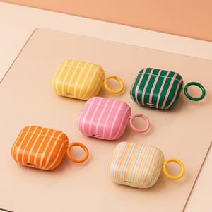 Könnte beinhalten: Fünf gestreifte AirPod-Hüllen in verschiedenen Farben: Gelb, Grün, Rosa, Orange und Pastell. Jede Hülle hat einen passenden Ring. Der Hintergrund ist hellpfirsichfarben. Schützende Accessoires.