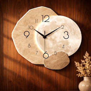 May include: A decorative wall clock with a unique design. The clock features a beige, stone-like frame with a circular moon-like face. Black hands indicate the time. The clock is mounted on a wooden wall.