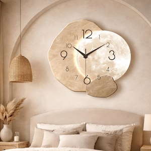 May include: A beige wall clock with a moon-like design. The clock features a large, circular face with a textured surface and black hands. The numbers are in a simple, modern font. The clock is mounted on a beige wall.
