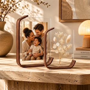 Könnte beinhalten: Zwei Fotorahmen aus Holz. Einer enthält ein Familienporträt, der andere Trockenblumen. Die Rahmen haben ein geschwungenes Design und sind aus dunklem Holz gefertigt. Die Displays stehen auf einer strukturierten, hellfarbigen Oberfläche.