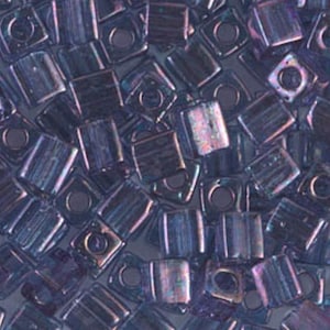 May include: Close-up of a pile of iridescent, square-shaped beads. The beads are a mix of dark blue and purple hues, with a subtle shimmer. Each bead has a square hole in the center, and they are scattered randomly.