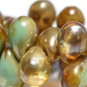 Pode incluir: Uma coleção de contas iridescentes em forma de gota, em tons de dourado e verde. As contas têm um acabamento brilhante, refletindo a luz e criando um efeito cintilante. As contas estão agrupadas, sugerindo que fazem parte de um projeto de artesanato ou design.