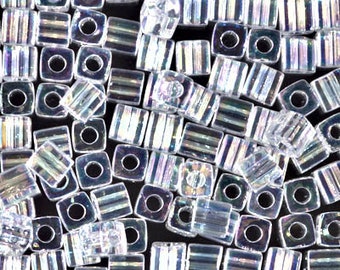 May include: A close-up of a pile of iridescent clear glass square beads. The beads have a faceted design and a hole in the center.