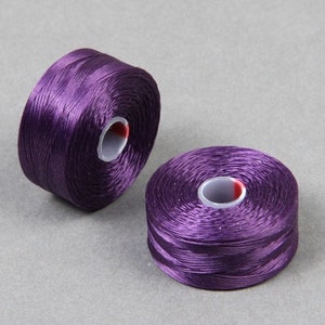 May include: Two spools of purple embroidery thread. The thread is wound around a white plastic spool with a red center.