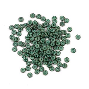 May include: A collection of small, teal-colored, flower-shaped beads scattered on a white surface. Each bead has a central hole surrounded by a detailed, textured design. The beads appear to be made of metal or a similar material.