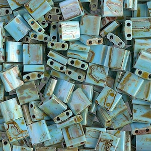 May include: A close-up of a pile of turquoise blue and brown glass tile beads. The beads are rectangular and have a mottled, iridescent finish.