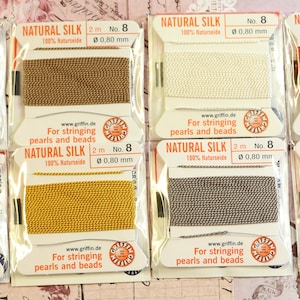 May include: Eight spools of natural silk thread for stringing beads. Each spool is labeled "Natural Silk" and has the length, number, and diameter printed on the label. The spools are arranged in two rows of four.