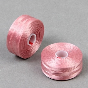 May include: Two spools of pink thread, each wound around a white plastic bobbin. The thread is smooth and shiny.