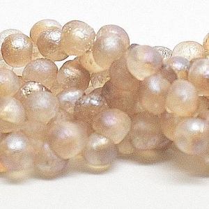 May include: Close-up of a strand of light brown, iridescent gemstone beads. The beads are round and clustered together, creating a textured surface. The beads have a pearlescent sheen, reflecting light.