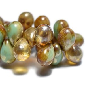 Pode incluir: Um fio de contas de vidro iridescentes em tons de verde e dourado. As contas são em forma de lágrima e têm um acabamento brilhante.