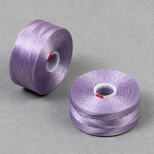 May include: Two spools of lavender-colored thread, each wound on a white plastic bobbin. The thread is smooth and shiny, and the bobbins have a red label.