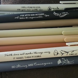 Puede incluir: Una variedad de bolígrafos en varios colores, incluyendo blanco, negro y tonos de marrón y gris. Cada bolígrafo tiene una cita inspiradora impresa, como "No temeré mal alguno" y "Sé fuerte y valiente."