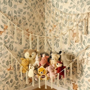 Op de afbeelding: Een crèmekleurige macramé speelgoedhangmat met een ruitvormig netpatroon en decoratieve franje-kwastjes langs de onderrand. De hangmat is in een hoek opgehangen en bevat een verzameling van diverse pluche knuffeldieren van verschillende groottes.