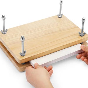 May include: A rectangular wooden flower press consisting of two light-colored wooden boards held together by four metal threaded rods with wing nuts at each corner, used for pressing flat items between the two wooden surfaces.