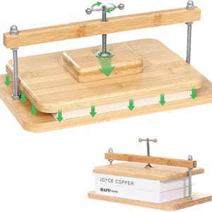 May include: A bamboo book press featuring two rectangular boards, a central metal screw mechanism with a handle, and two threaded metal rods with nuts. The device is shown compressing two white books labeled JOYCE COPPER and HAPPINESS.