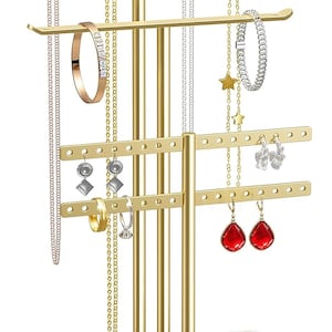 May include: A gold-toned metal jewelry organizer stand with multiple tiers for necklaces, bracelets, and earrings. The stand features a white rectangular base tray, horizontal bars with holes for earrings, and vertical rods for hanging items.