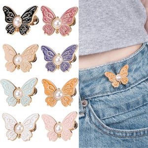 May include: A collection of eight butterfly-shaped pins in black, pink, beige, purple, light blue, orange, white, and light pink, each featuring a central white faux pearl and intricate filigree detailing, shown attached to blue denim jeans.