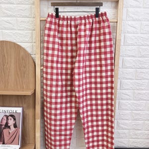 Pu&ograve; includere: Pantaloni a quadri rossi e bianchi appesi a un attaccapanni di legno. I pantaloni hanno un elastico in vita e un design a gamba larga. Il tessuto sembra leggero, adatto all'abbigliamento casual. Il motivo &egrave; un classico quadretto vichy.