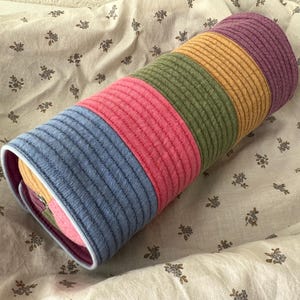 May include: A colorful, cylindrical pillow with a striped design. The pillow features bands of blue, pink, green, yellow, and purple. The pillow is resting on a floral patterned fabric.